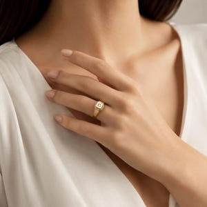 May include: A gold signet ring featuring a square-shaped face with a central letter D. The ring has a polished metallic finish and a smooth band, showcasing a clean design that sits on the finger with a classic and understated appearance.