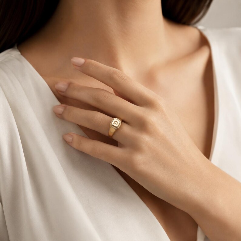 May include: A gold signet ring featuring a square-shaped face with a central letter D. The ring has a polished metallic finish and a smooth band, showcasing a clean design that sits on the finger with a classic and understated appearance.