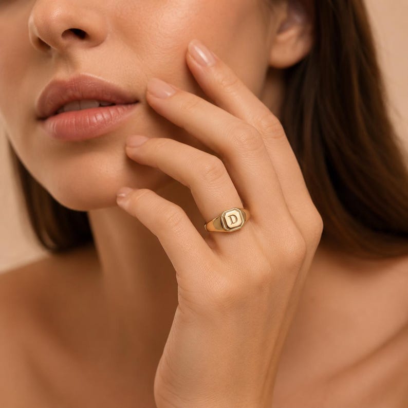 May include: A gold signet ring featuring an octagonal face with the letter D engraved in the centre. The ring has a polished finish and a smooth, tapered band that sits comfortably on the finger, showcasing a classic and clean design aesthetic.