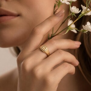 May include: A gold signet ring featuring an octagonal face with the letter D engraved in the centre. The ring has a polished finish and a smooth band, showcasing a classic design that sits on the finger with a clean and refined appearance.