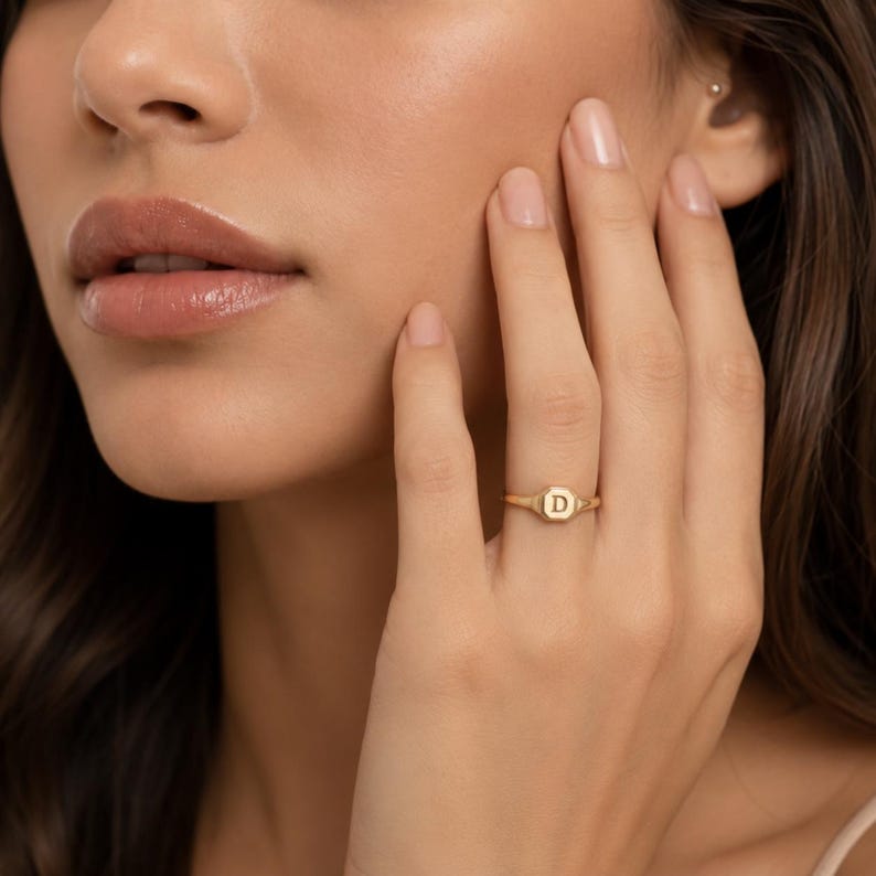 May include: A gold signet ring featuring a small, flat, octagonal face engraved with the capital letter D. The ring has a thin, polished band and is worn on the ring finger, showcasing a metallic finish and a simple, clean design.