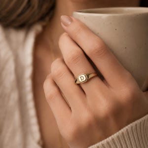 May include: A gold signet ring featuring an octagonal face with the letter D engraved in the centre. The ring has a polished finish and a smooth, tapered band that sits on the finger, showcasing a classic design with clean, defined edges.