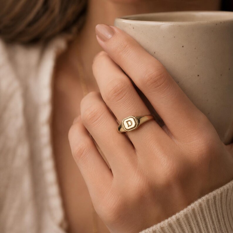 May include: A gold signet ring featuring an octagonal face with the letter D engraved in the centre. The ring has a polished finish and a smooth, tapered band that sits on the finger, showcasing a classic design with clean, defined edges.