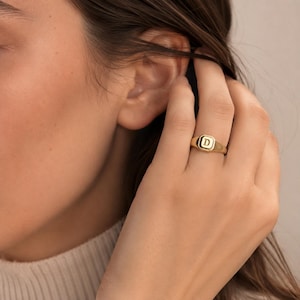May include: A gold signet ring featuring a square-shaped face with a beveled edge and the letter D engraved in the centre. The ring has a polished finish and a smooth, tapered band that sits comfortably on the finger, showcasing a classic design.