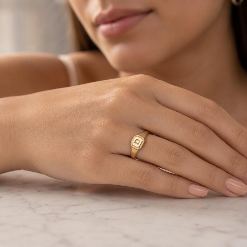 May include: A gold signet ring featuring a square face with a recessed border and the letter D engraved in the centre. The ring has a polished finish and a slim band that tapers towards the back, showcasing a classic design with clean lines.