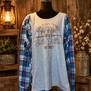 Könnte beinhalten: Ein Langarmshirt mit einem mittelgrauen Bereich und Ärmeln mit blau-weiß-rotem Karomuster. Auf der Vorderseite befindet sich ein brauner Schriftzug: Why y'all trying to test the Beth Dutton in me? mit einer kleinen Berggrafik darüber.