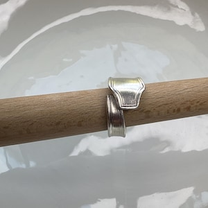 May include: A silver-toned ring crafted from the handle of a metal spoon, featuring a bypass design where the two ends overlap. The ring is displayed wrapped around a smooth, light-colored wooden dowel, showcasing its metallic finish and curved shape.