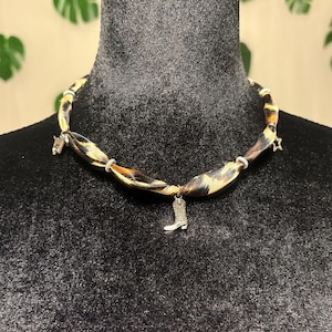 May include: A choker necklace featuring a twisted fabric band with a brown and black leopard print pattern. The necklace is accented with small silver-toned metal charms, including a central cowboy boot, a star, and other small shapes.