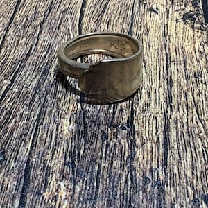 May include: A silver-toned metal ring fashioned from a repurposed spoon handle. The band features a smooth, curved surface with visible stamped text on the interior that reads ONEIDA LTD. The metal has a polished finish with subtle signs of wear.