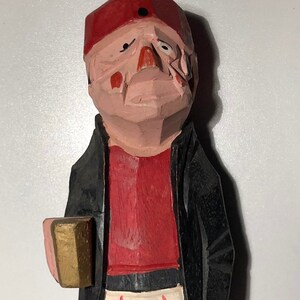 Op de afbeelding: Een houten beeldje van een honkballer met een rode pet, een rood shirt, een witte broek met rode strepen en een zwart jack. Het beeldje houdt een klein houten voorwerp in zijn linkerhand en heeft een houten houten been.