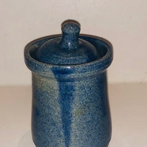 Peut inclure: Un petit pot en céramique bleu avec un couvercle. Le pot a un glaçage moucheté et une bande bleu clair sur le côté. Le couvercle est un simple bouton.