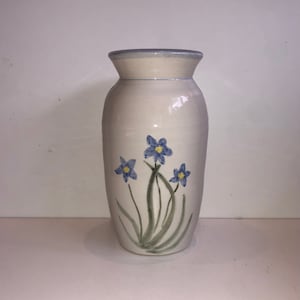以下が含まれることがあります： 淡い青色の縁と、緑の茎を持つ3つの青い花のイラストが描かれた白いセラミック花瓶。花瓶はわずかにフレアした上部と滑らかな円筒形のボディを持っています。花は手描きです。