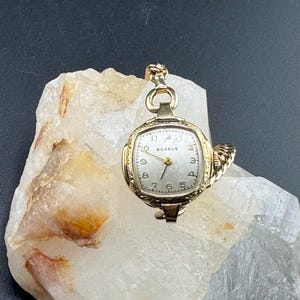 May include: A vintage gold-toned wristwatch with a square face and a light-colored dial. The watch has a delicate design with ornate detailing around the face and a matching chain. The watch face displays the brand name "Behru" and the numbers 1 through 12.