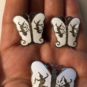 May include: A set of three silver butterfly earrings with white enamel and black silhouette designs. The butterflies have intricate details and are a unique and elegant piece of jewelry.