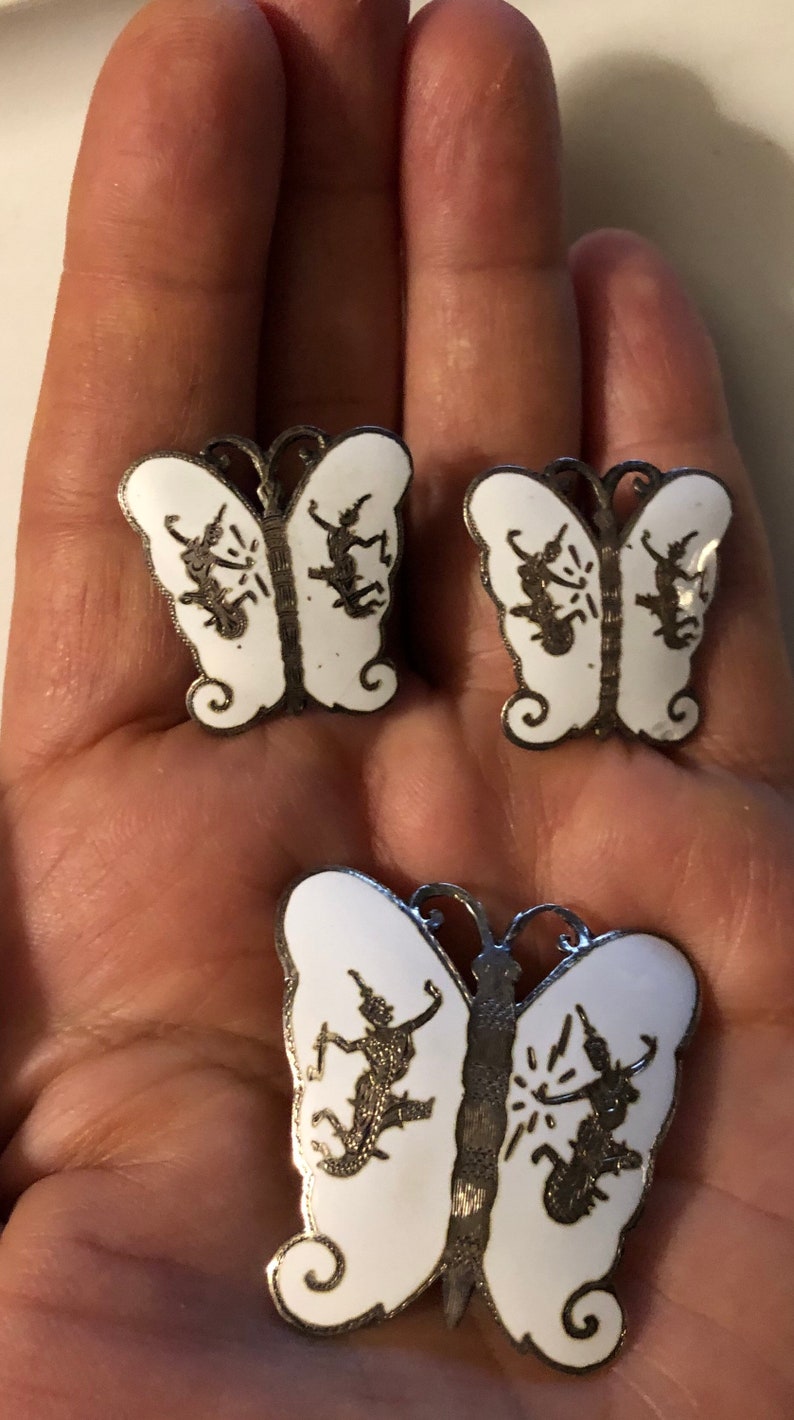 May include: A set of three silver butterfly earrings with white enamel and black silhouette designs. The butterflies have intricate details and are a unique and elegant piece of jewelry.
