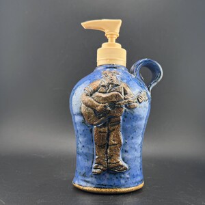 May include: Blue ceramic soap dispenser with a brown relief of a person playing a guitar. The dispenser has a beige pump and a small handle. The ceramic has a speckled texture.