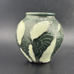 Puede incluir: Un pequeño jarrón de cerámica redondo con un fondo verde oscuro y un patrón de formas de hojas verde claro. El jarrón tiene un borde ligeramente ensanchado y un acabado liso y brillante. El diseño recuerda a la naturaleza.