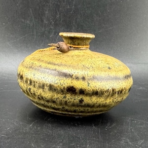 Op de afbeelding: Een handgemaakte, ronde keramische vaas met een smalle hals en een getextureerd, gespikkeld oppervlak. De vaas is voornamelijk geel met donkerbruine horizontale banden en accenten. Een touw en metalen sluiting bevestigen het deksel.