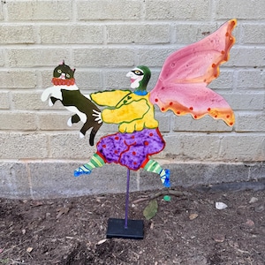 May include: A whimsical metal garden stake featuring a fairy-like figure holding a black and white cat. The fairy has pink wings, a yellow top, purple polka-dot skirt, and green and blue accents. The stake is mounted on a black base.