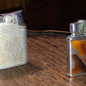 May include: Two vintage silver lighters. One has a textured, brick-like pattern. The other has a brown and gold design with a map-like pattern.