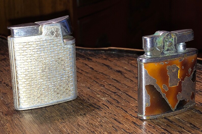 May include: Two vintage silver lighters. One has a textured, brick-like pattern. The other has a brown and gold design with a map-like pattern.