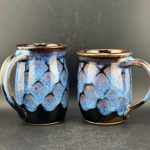 Puede incluir: Dos tazas de cerámica con un esmalte brillante que presenta un patrón de escamas en azul y negro. Cada taza tiene un asa curva y un borde marrón oscuro. La base es de color negro sólido, transformándose en un diseño azul y negro texturizado.