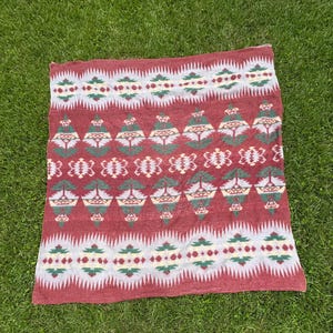 Puede incluir: Una manta tejida rectangular con un color base rojo intenso y patrones geométricos repetitivos en tonos crema, verde bosque y amarillo apagado. El diseño incluye bandas horizontales de triángulos dentados y motivos florales.