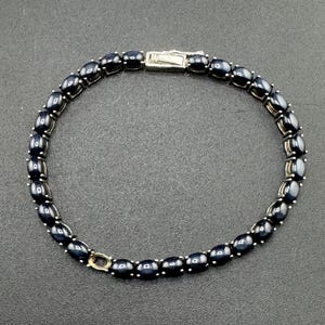 Op de afbeelding: Een armband met donkerblauwe, ovale edelstenen. De armband heeft een zilverkleurige sluiting en is geplaatst tegen een donkere achtergrond. De edelstenen zijn gelijkmatig verdeeld en verbonden door zilverkleurig metaal.
