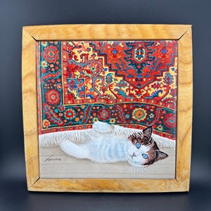 May include: A framed painting of a white cat with blue eyes lying on a red, green, and yellow patterned rug. The rug has fringe and the cat is looking directly at the viewer.