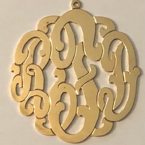 Puede incluir: Un colgante de monograma de metal dorado con un diseño decorativo y adornado. El colgante presenta tres letras entrelazadas.