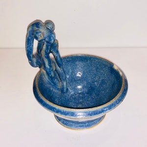 Puede incluir: Un pequeño cuenco de cerámica azul con un asa única con forma de figura estilizada. El cuenco tiene un esmalte moteado y un borde ligeramente elevado.