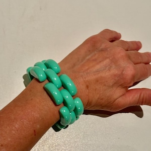 Puede incluir: Un brazalete de plástico turquesa con un diseño de cadena enlazada. El brazalete se lleva en la muñeca de una persona.