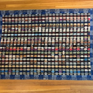 Può includere: Una trapunta vintage con un bordo blu e un motivo ripetitivo di quadrati di tessuto colorati. La trapunta è realizzata con diversi tessuti, tra cui cotone e lino.