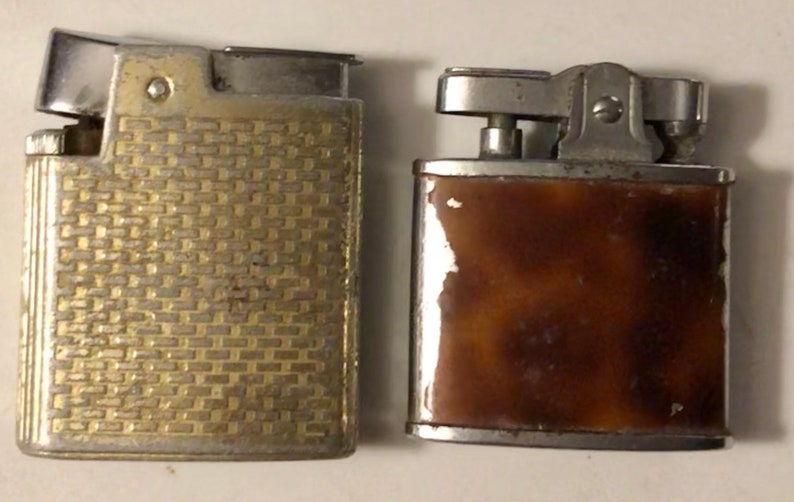 May include: Two vintage lighters, one with a gold-toned, textured finish and the other with a brown, mottled finish.
