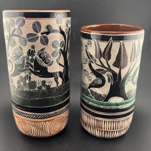 Może przedstawiać: Dwa ceramiczne wazony z ręcznie malowanymi wzorami. Wazony mają czarne motywy drzew i zwierząt na beżowym tle. Obręcze i podstawy mają dekoracyjne pasy w kolorze czarnym, brązowym i białym. Wazony są cylindryczne i mają wysokość około 20 cm.