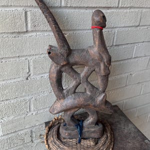 Puede incluir: Escultura de madera tallada de color marrón oscuro. Presenta una figura humana estilizada con cuello largo y forma de ave. Una banda roja rodea el cuello. La escultura está montada sobre una base tejida.