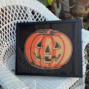 May include: A framed Halloween decoration featuring a smiling jack-o'-lantern. The pumpkin is orange with a brown paper background and black facial features. The frame is black, and the artwork is displayed on a white wicker chair.