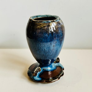 May include: A small, blue and brown ceramic vase with a scalloped base and gold accents. The vase has a glossy finish and a unique, hand-crafted look.