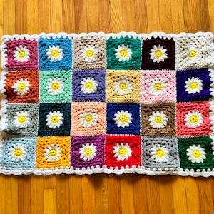 Puede incluir: Una manta de crochet colorida con un patrón de margarita. La manta está hecha de cuadrados de varios colores, incluyendo rosa, azul, verde, amarillo y marrón. Cada cuadrado presenta una margarita blanca con un centro amarillo.