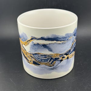 May include: A cylindrical ceramic container with a cream-colored interior and rim. The exterior features a watercolor design of blue and gold abstract mountain-like shapes. The base is a pale lavender color.