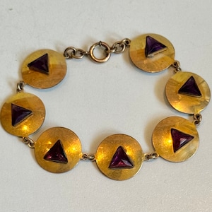 May include: A gold-toned bracelet featuring circular links, each adorned with a triangular, deep purple gemstone. The bracelet is connected by small, delicate links and has a clasp closure. The design is simple yet elegant.