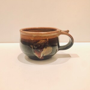 Puede incluir: Una taza de cerámica pequeña y marrón con un patrón de esmalte único. La taza tiene un cuerpo redondeado y un asa ligeramente descentrada.
