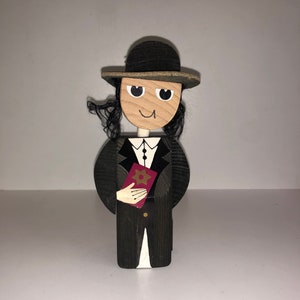 Könnte beinhalten: Holzfigur einer Person, die einen schwarzen Hut, einen schwarzen Anzug trägt und ein rotes Buch mit einem goldenen Davidstern auf dem Einband hält.
