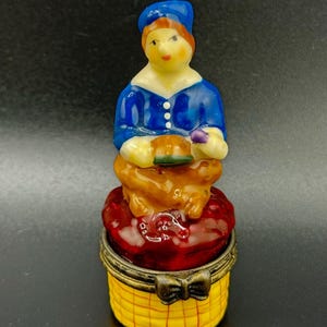 Puede incluir: Una caja de porcelana para baratijas con una figurita pintada a mano de una persona que lleva un sombrero y una chaqueta azules, sentada en un cojín rojo con una base a cuadros amarillos y marrones.