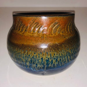Puede incluir: Una pequeña maceta redonda de cerámica con un esmalte marrón, naranja y azul. La maceta tiene un diseño texturizado alrededor del borde superior.