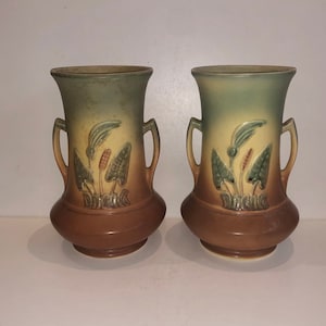 Könnte beinhalten: Zwei Keramikvasen mit einem Design mit zwei Griffen. Die Vasen weisen einen Farbverlauf von Grün über Beige bis Braun auf. Jede Vase hat ein botanisches Reliefdesign aus Blättern und Blumen.