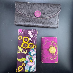 Könnte beinhalten: Eine dunkelbraune, texturierte Ledergeldbörse mit lila Blumenknopfverschluss, ein hohes, rechteckiges, lila Stoffetui mit gelb-blauem Blumenmuster und eine kleine, rechteckige, lila Stoffgeldbörse mit einem gelben, runden Knopf.