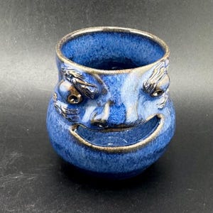 May include: A handmade ceramic mug with a smiling face design. The mug is glazed in shades of blue with brown accents around the eyes, nose, and mouth. The mug has a unique, textured appearance and is suitable for beverages.