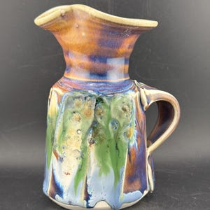 May include: A ceramic pitcher featuring a flared spout and a side handle. The exterior displays a multi-colored glaze with vertical drips in shades of green, blue, and brown, characterized by a visible crackle finish throughout the surface.