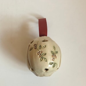 Puede incluir: Un adorno de cerámica blanca con forma de bola con una cinta roja atada. El adorno está decorado con un diseño floral marrón y verde.